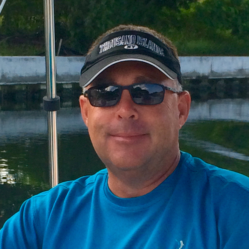 a man in a blue shirt and black visor smiles while standing on a boat