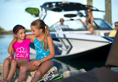 two girls sitting on rocky beach in front of boats with arms around each others shoulders