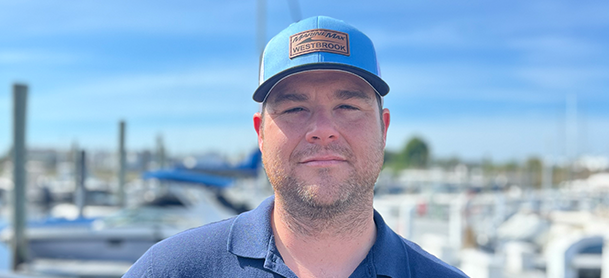 Robert Finch wearing a blue cap with text and a navy polo shirt standing by a sunny marina.