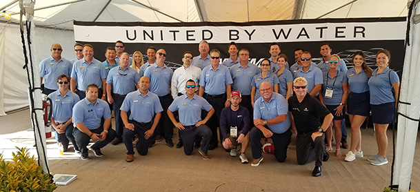 A group of MarineMax employees in light blue polo shirts lined up for a photo in front of a sign that says "United by Water"
