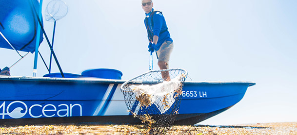 A man in a blue shirt standing on a blue boat, holding a net filled with trash on the surface of the beach