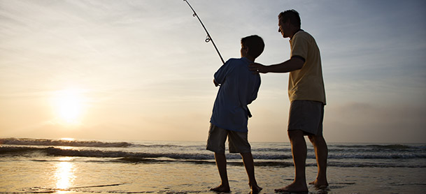 A man and boy fishing in shallow water while standing on sand as the sun sets