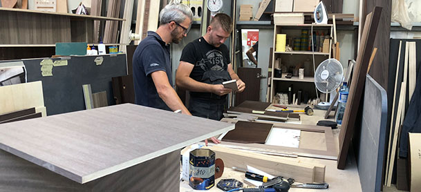 Two men working on wooden boards in a factory, with various tools around them