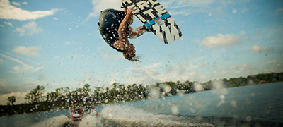 Male upside down in the air doing a wakesurfing trick with water splashing and a boat in the background