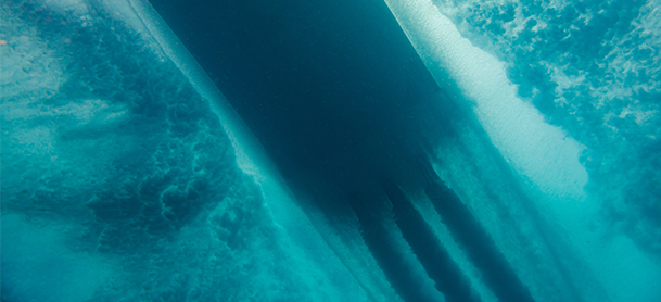 Underwater view of a ship’s hull with bubbles and turbulence in the surrounding blue water.