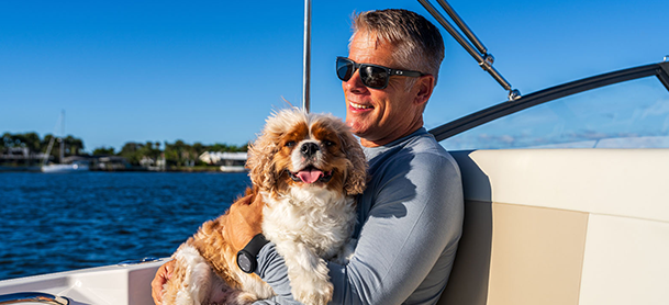 A man wearing sunglasses smiles while holding a fluffy dog on a boat.