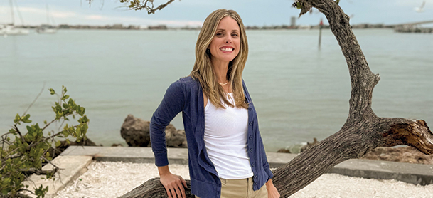 Bridget Pinsonneault, in a blue cardigan and white shirt leans on a twisted tree branch by a calm waterfront.