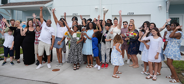 A joyful, diverse group stands in front of houses, hands raised in celebration.