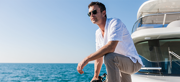 A man in a white shirt and sunglasses leans on a yacht railing, gazing at the ocean under a clear blue sky.
