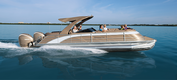 A tan and silver luxury pontoon boat cruises swiftly on clear blue water under a sunny sky.