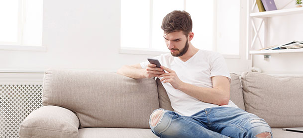 A man in a white shirt and jeans sitting on a couch looking at a smart phone