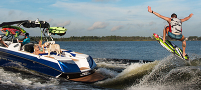 male wakesurfer doing a trick with arms in the air behind a boat with people on aft deck seating