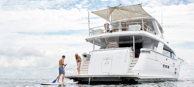 Male on stand up paddle board behind a large yacht with a woman standing on the swim platform