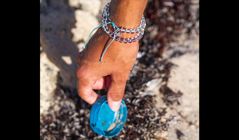 a hand picking up a bottle cap off of the beach while wearing a colorful 4ocean bracelet