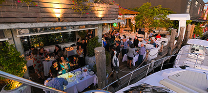 a view of people gathered at a boating event with people standing and seated at tables with food and drinks