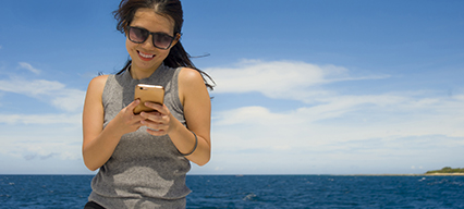 a woman holding a smartphone and smiling with blue water and clear skies behind hera woman holding a smartphone and smiling with blue water and clear skies behind her