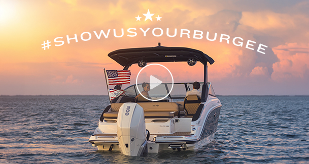 A boat with an American flag cruises on calm water under a vibrant sunset sky. The text "#ShowUsYourBurgee" arcs above, creating a serene and adventurous mood.