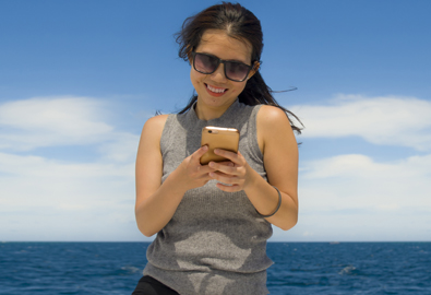 Smiling woman looking at phone through sunglasses on a beautiful sunny day by the ocean.