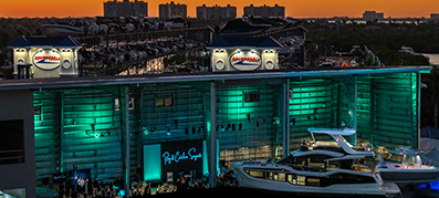 A large marina building illuminated with teal lights at dusk displaying luxurious yachts.