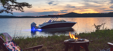 Cozy lakeside scene at sunset with a boat on the water, two chairs near a small campfire, and trees framing the view, evoking tranquility.