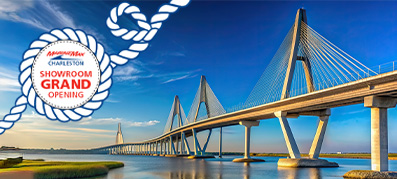 Suspension bridge over a calm river under a bright blue sky, reflecting in the water. Text in rope border reads "Charleston Showroom Grand Opening."