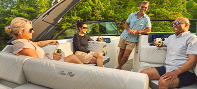 Four people relax and enjoy drinks on a luxury boat. They are seated on white, cushioned benches with "Sea Ray" branding. The mood is cheerful and relaxed, surrounded by lush green trees.