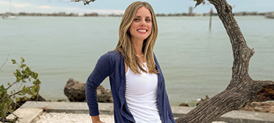 Bridget Pinsonneault, in a blue cardigan and white shirt leans on a twisted tree branch by a calm waterfront.