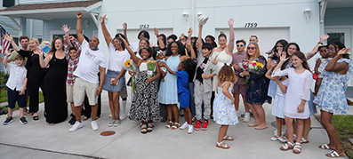 A joyful, diverse group stands in front of houses, hands raised in celebration.