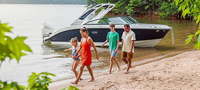 A family of four walks along a sandy beach by a lake, with a boat docked nearby.