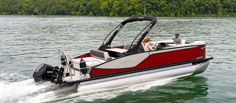 A red and gray Harris pontoon with two people is cruising on a calm lake.