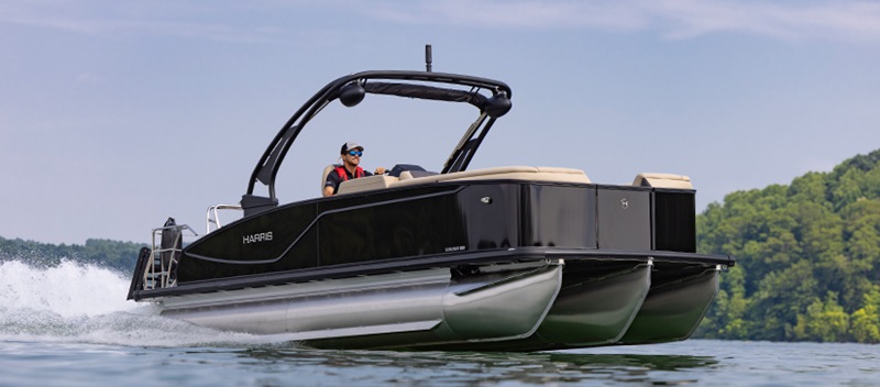 A sleek black Harris pontoon speeds across a calm lake, with a person wearing a life jacket steering.