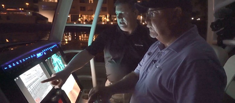 A man in a black polo pointing at a navigation screen on a boat while a man in a blue polo looks ahead and drives