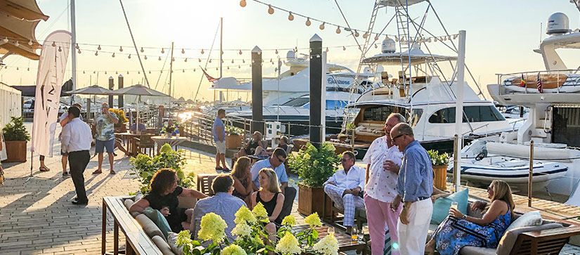 A group of people chatting on a dock with yachts beside them, as the sun sets in the background
