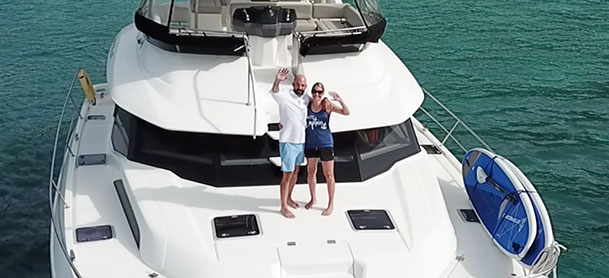 A couple stands on the deck of a white yacht, smiling and waving.