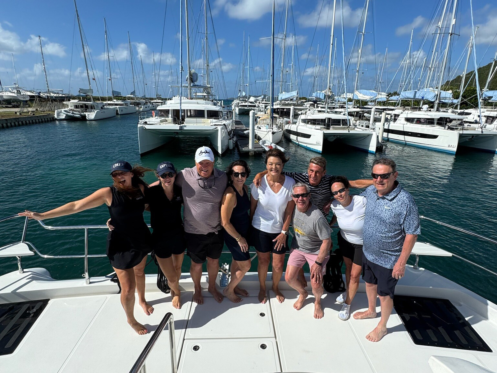 Group of people posing on a yacht docked in a marina with sailboats in the background.