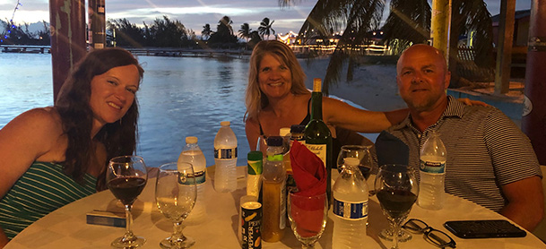 A man and two women seated at a dinner table, with the sun setting over the water behind them