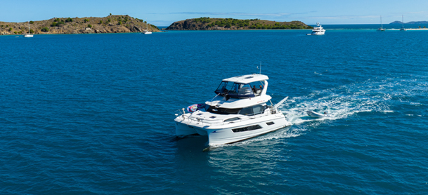 A white catamaran cruises through calm blue waters, leaving a trail of gentle waves. In the background, small islands and boats dot the sunlit horizon.