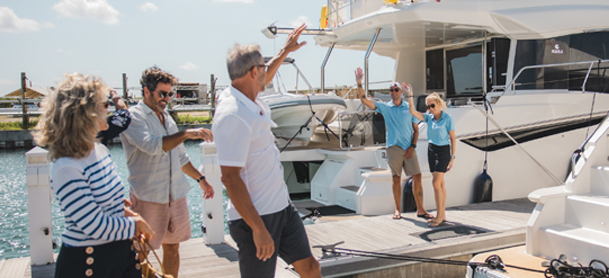 A group of people on a sunny dock, smiling and waving at a yacht. The scene conveys a sense of joy and camaraderie near the water.