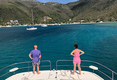 A man and woman standing on the stern of a power catamaran, looking toward the coast of the British Virgin Islands