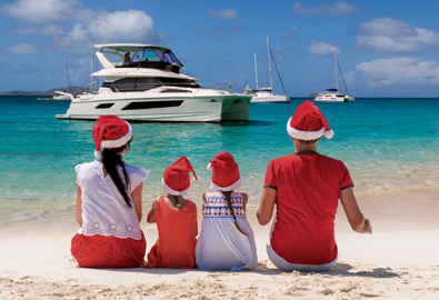 Family sitting on beach with santa hats looking out to water where power catamaran is anchored.