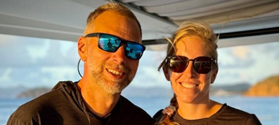 a smiling man and woman wearing sunglasses on a boat surrounded by a serene ocean backdrop and softly clouded sky