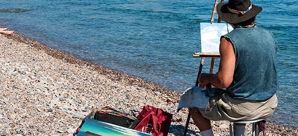 A man sits on a rocky beach and paints the ocean in front of him