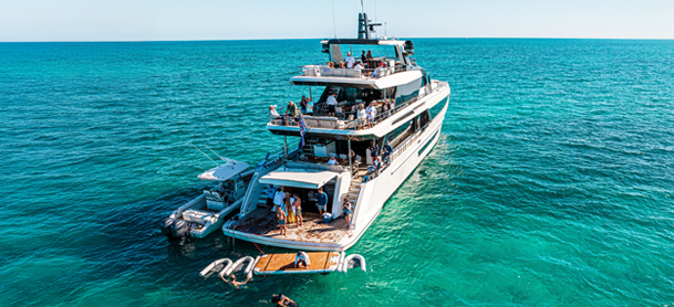 A luxury yacht floating on clear turquoise water, with several people on deck enjoying the sunny day.