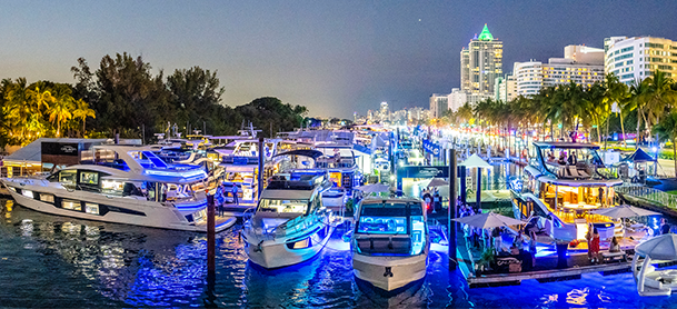 Brightly lit marina at night with numerous yachts docked; city skyline and palm trees in the background.