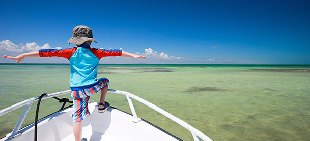 A boy wearing a hat standing at the stern of a boat, looking out into the water