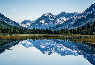 A scenic view of a mountain range in Alaska next to a lake, with grass and trees in between
